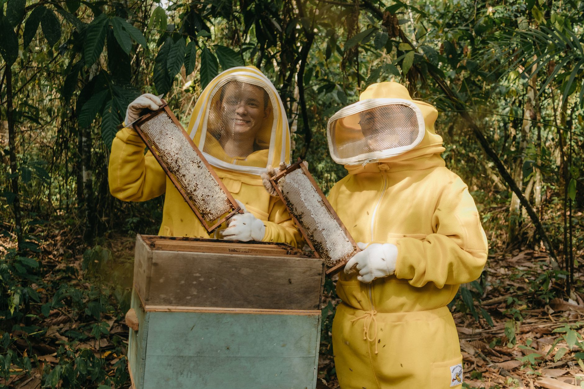 a child in a white beekeeping suit holding a frame while an adult supervises